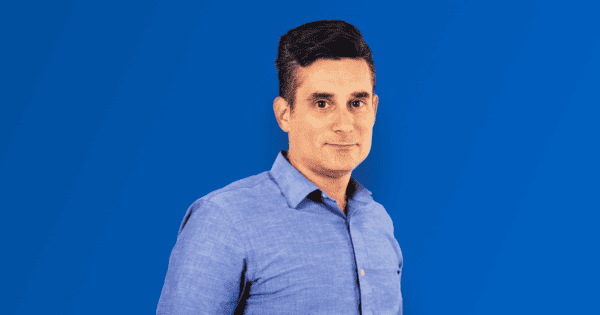 Steve Shaner in blue button down shirt standing with hands in pockets with a blue gradient background.