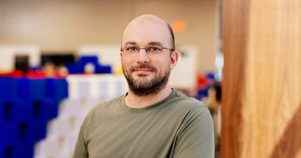A bearded man with glasses stands indoors, smiling slightly at the camera. He is wearing a casual green t-shirt. The background is softly blurred, featuring large, colorful blocks and a wooden partition, suggesting a creative or playful environment.