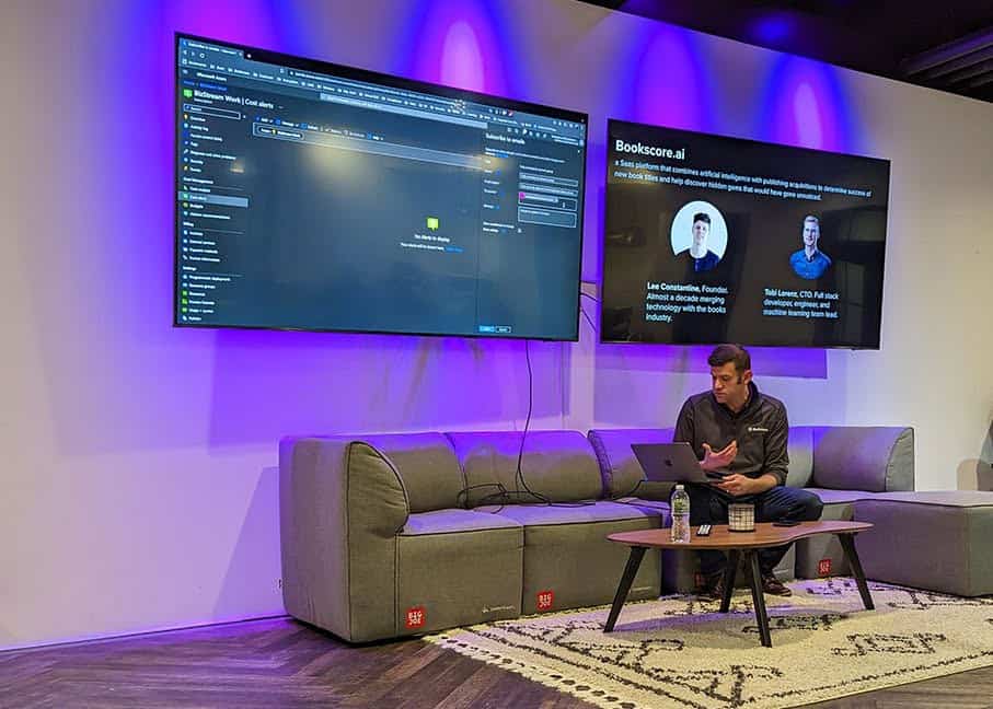 Photo of a modern conference or presentation setup with a man seated on a stage with two large screens behind him. The screens display code and presentation slides in a dimly lit room with purple accent lighting. The scene suggests a tech talk or workshop setting.