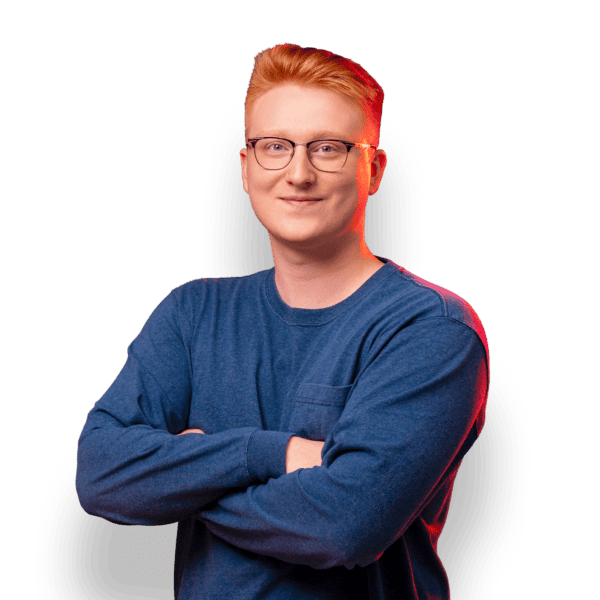 A young man with red hair and glasses is standing against a black background with red lighting. He is wearing a blue long-sleeve shirt and has a relaxed, confident expression. His arms are crossed in front of him.