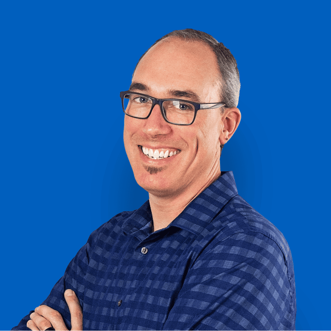 Headshot of Drew Veach, smiling and wearing glasses with a blue plaid shirt, set against a solid blue background.