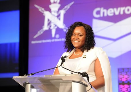 A woman in a white outfit stands at a podium, smiling while delivering a speech at an event co-hosted by NSBE and Chevron, with event branding visible behind her.