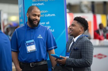 Two men, one in a blue polo shirt and the other in a plaid suit jacket, chat and laugh while networking in front of a corporate booth at a professional event.
