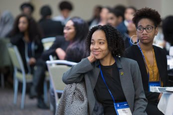 A young woman in professional attire smiles while seated at a conference roundtable surrounded by other attendees, wearing a blue lanyard and name badge.