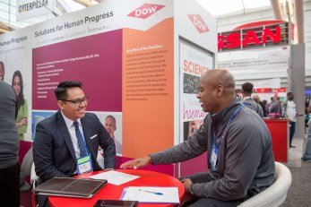 Two men engage in conversation at a DOW recruitment or career fair booth, seated at a small red table with brochures and materials, in a large expo hall.