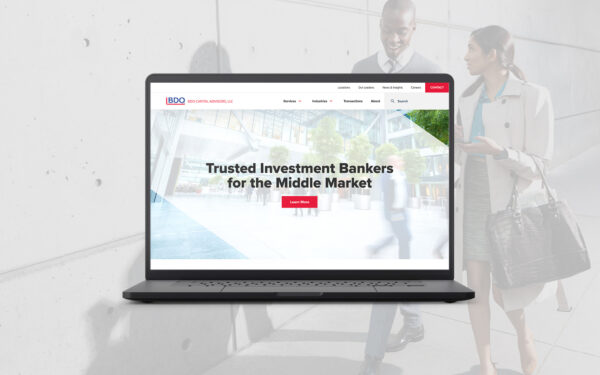 A laptop displaying the BDO Capital Advisors website homepage, featuring a banner with the headline "Trusted Investment Bankers for the Middle Market" and a red "Learn More" button. The navigation menu at the top includes links for Services, Industries, Transactions, About, and more. The background image of the banner shows a modern office building with glass windows and trees, creating a professional atmosphere. In the background outside of the laptop frame, a man and a woman are engaged in conversation, adding a sense of business context to the image.
