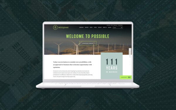 A laptop displaying the Moss Adams website on its screen, with the Moss Adams logo and navigation menu at the top. The website features a hero banner with the text "WELCOME TO POSSIBLE" above an image of wind turbines in a scenic landscape. Below the banner, there's a section highlighting "111 YEARS IN BUSINESS" in large text. The overall theme of the website conveys a professional and optimistic approach, using a combination of green and white colors. The laptop is set against an aerial view of an urban intersection with a dark, textured overlay.