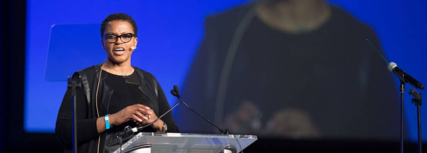 A woman in black attire and glasses speaks confidently at a clear podium during a large conference, with a microphone and blue stage backdrop behind her.