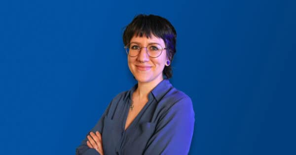 Person with short dark hair, wearing glasses and a blue shirt, standing confidently with arms crossed against a solid blue background, smiling warmly.