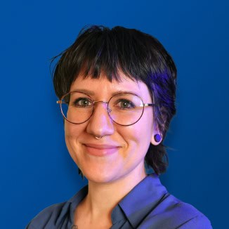 Person with short dark hair, wearing glasses and a blue shirt, standing confidently with arms crossed against a solid blue background, smiling warmly.