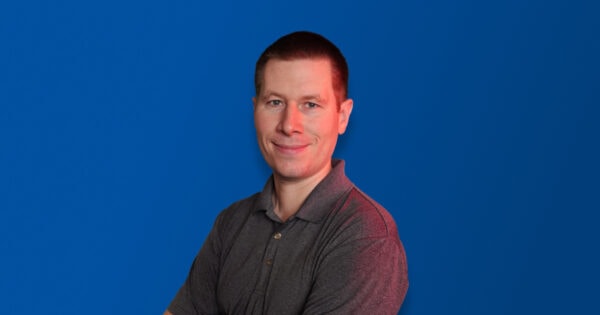 Person with short dark hair, wearing a gray polo shirt, standing confidently with arms crossed against a solid blue background, with a subtle red lighting effect on one side, smiling warmly.