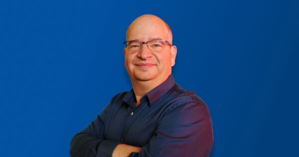 Person with glasses and a shaved head, wearing a dark blue shirt, standing confidently with arms crossed against a solid blue background, smiling warmly.
