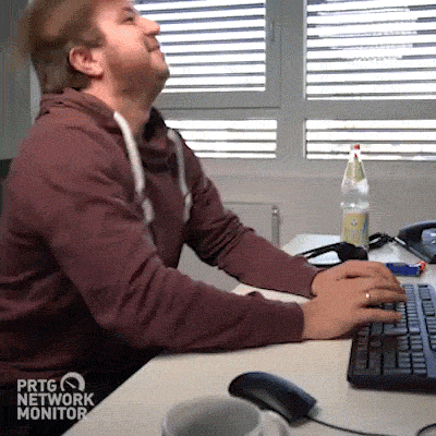 Frustrated man in a hoodie slams his keyboard repeatedly at a desk, then throws his head back in exasperation. “PRTG Network Monitor” logo visible in the corner.