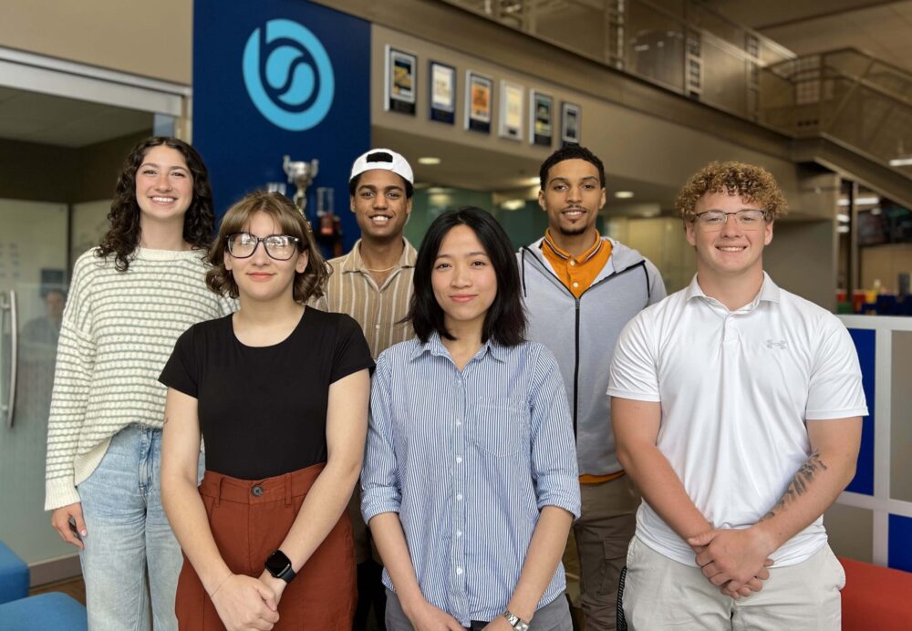 Group photo of six smiling young adults standing inside an office space with the BizStream logo on the wall behind them