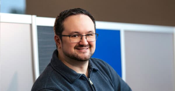 Kyle Dhyne smiling in glasses and a dark zip-up sweater, seated at his workstation in the BizStream office.