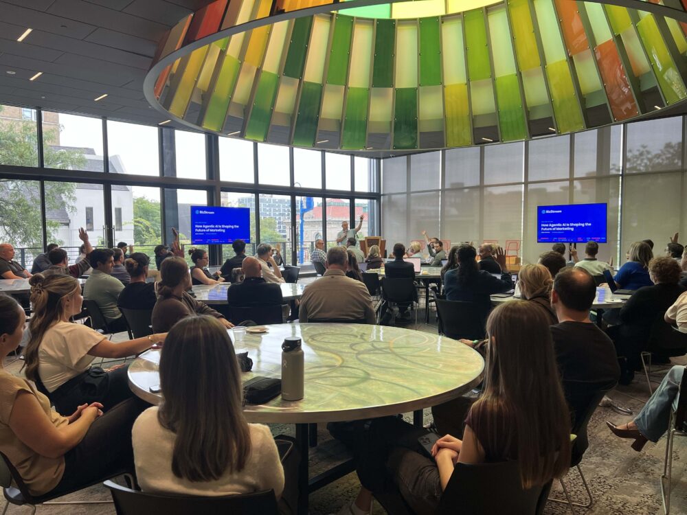 Brian McKeiver presents to a full, engaged audience with several hands raised during BizStream's agentic AI session at Tech Week Grand Rapids, beneath the colorful glass SkyCone at GRCC's Secchia Piazza.