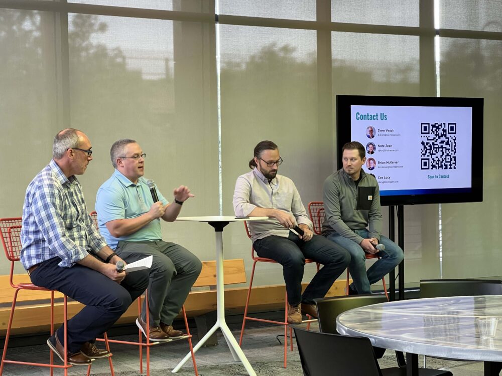 Four BizStream panelists seated on stools take audience questions beside a "Contact Us" slide, with one speaking into a microphone, during the Q&A at Tech Week Grand Rapids.