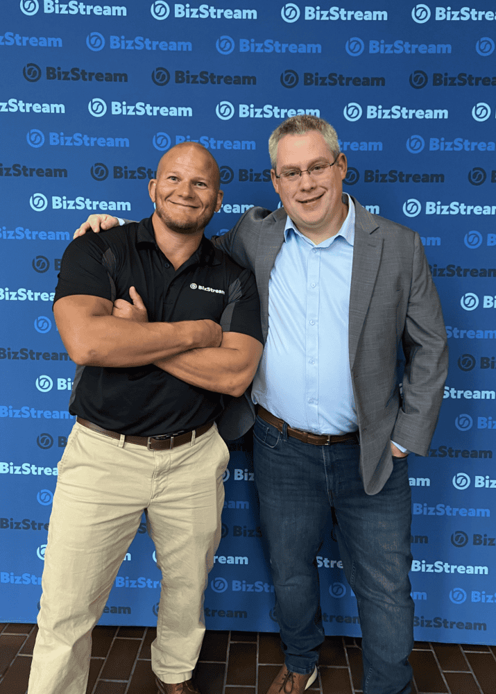 BizStream Founder Mark Schmidt and Co-owner Brian McKeiver pose in front of a BizStream logo backdrop at the 25th anniversary celebration.
