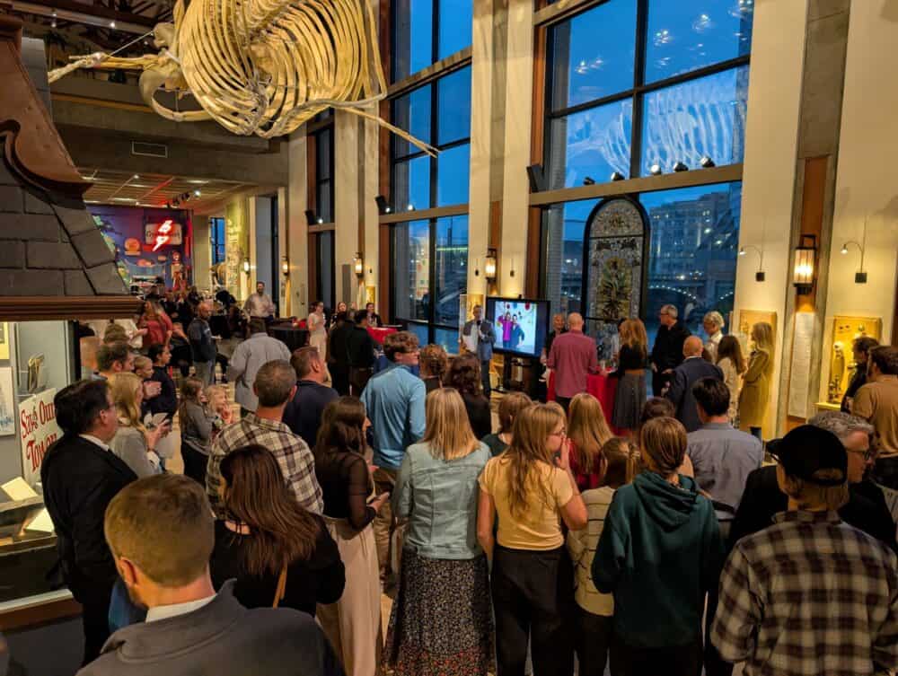 Guests gather beneath a whale skeleton and tall windows overlooking downtown Grand Rapids during the anniversary toast.