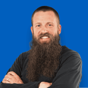 Headshot of Josh Trench, smiling with a long beard, wearing a black shirt, set against a solid blue background.